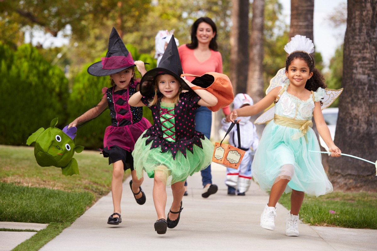 three kids trick or treating with mom behind them on sidewalk